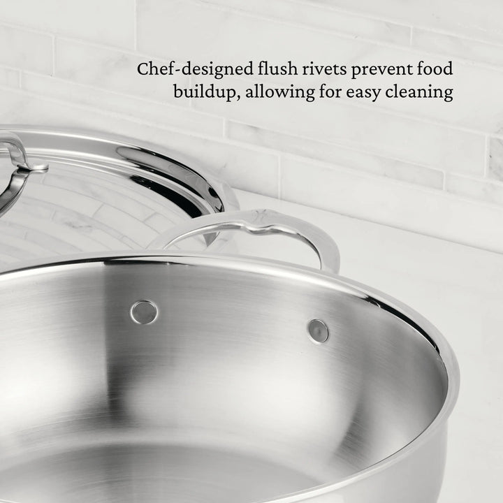 A ProBond® Luxe™ Professional Clad Stainless Steel Essential Pan with Lid sits on a white countertop. Chef-designed, flush rivets inspired by Italian craftsmanship prevent food buildup for effortless maintenance and easy cleaning.
