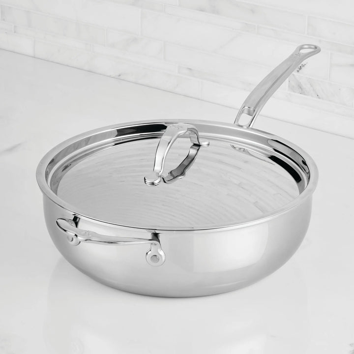 A ProBond® Luxe™ Professional Clad Stainless Steel Essential Pan with Lid and dual handles rests on a white marble counter, set against a white tiled backsplash and showcasing premium Italian-inspired craftsmanship.