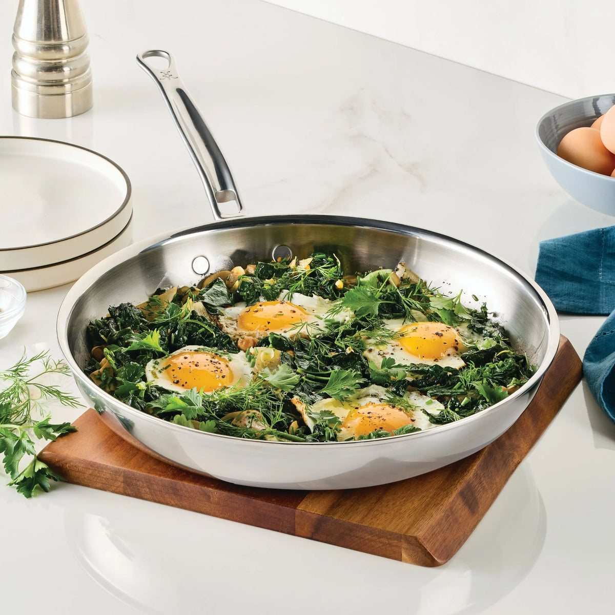 A ProBond® Luxe™ Polished Clad Stainless Steel Skillet sits on a wooden board holding cooked eggs with greens and herbs. Nearby on the white countertop are plates, a pepper grinder, a cloth napkin, and a bowl of eggs.