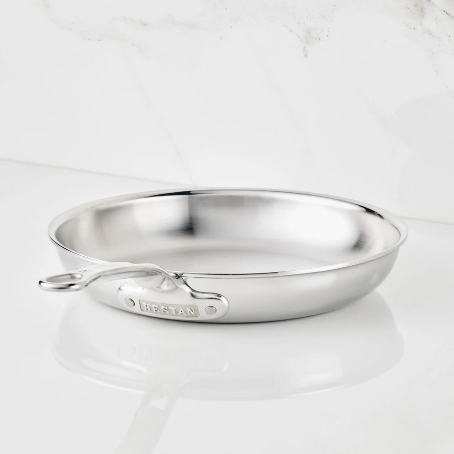 A ProBond® Luxe™ Polished Clad Stainless Steel Skillet with a riveted handle sits on a white surface against marble. “ProBond® Luxe™” is engraved on the handle, highlighting premium construction and shine.