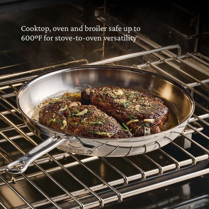 A ProBond® Luxe™ Polished Clad Stainless Steel Skillet with two seared steaks rests on an oven rack, demonstrating its versatility—its safe for cooktop, oven, and broiler use up to 600°F.
