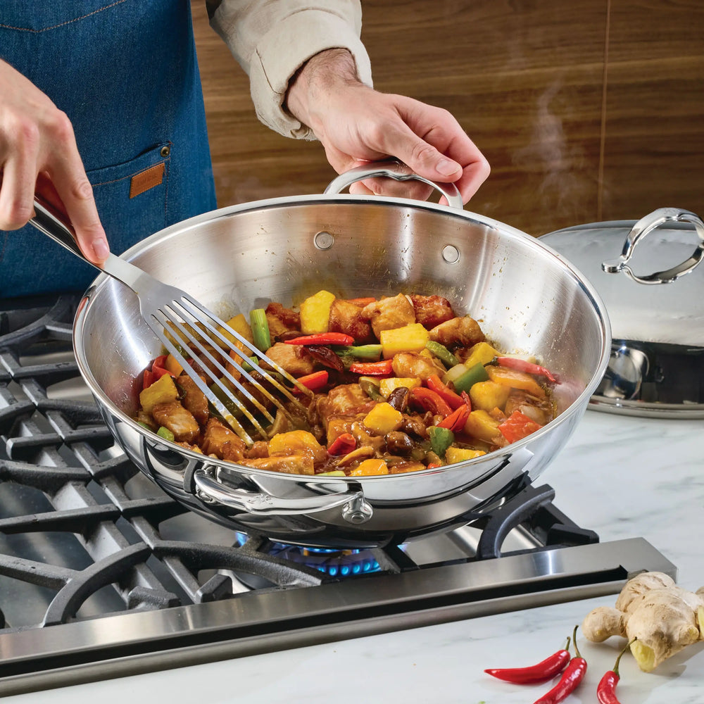 Wearing a blue apron, a person stirs vibrant meat, peppers, pineapple, and sauce with a metal spatula in a 14-inch ProBond® Luxe™ Polished Clad Stainless Steel Wok with Dome Lid on a gas stove. Ginger and red chilies are on the counter.