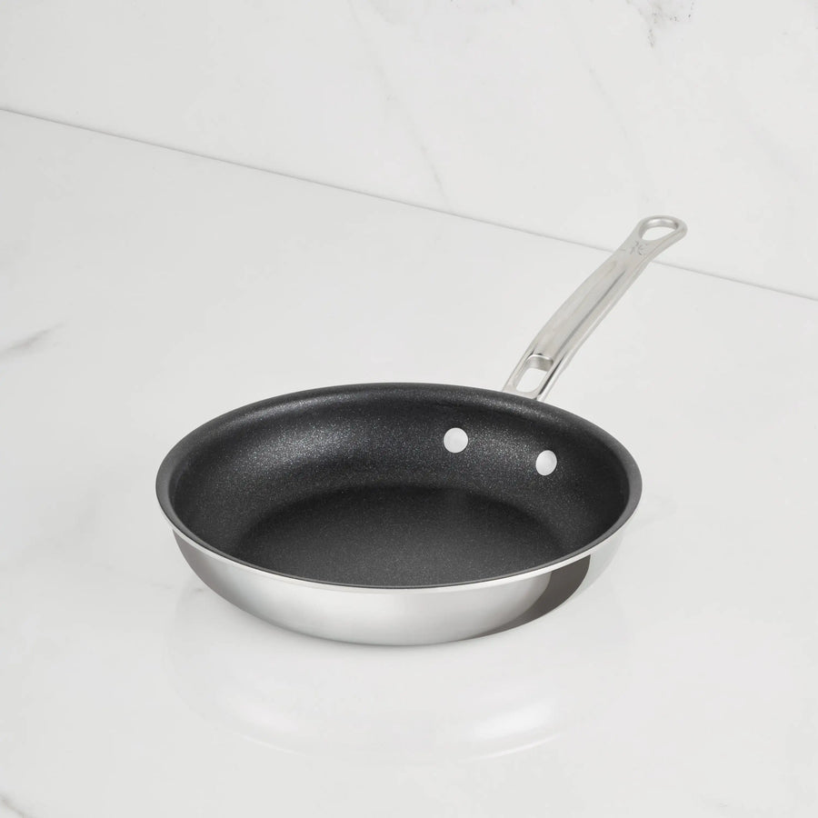 A ProBond Luxe® Polished Clad Stainless Steel Skillet with TITUM® Nonstick, featuring a sleek metal handle and crafted with exquisite Italian craftsmanship, sits on a white marble surface with a white marble background.