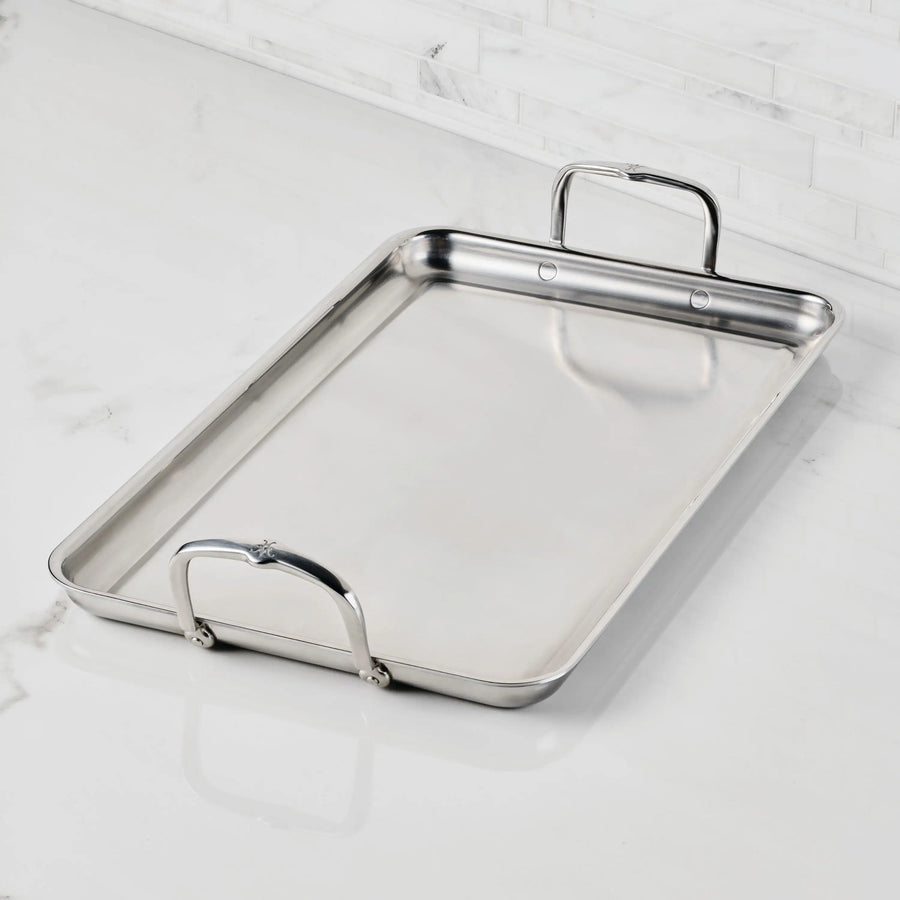 The Hestan Provisions Double Burner Griddle (21x13) features raised edges and two large handles, shown resting on a white marble countertop with a white tiled backsplash in the background.