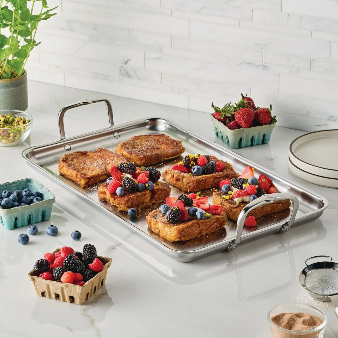 A stainless steel tray from the NanoBond Titanium x Tyler Florence Cookware Set, 41-Piece sits on a kitchen counter with French toast topped with mixed berries. Around it are berry cartons, pistachios, potted herbs, and stacked plates.