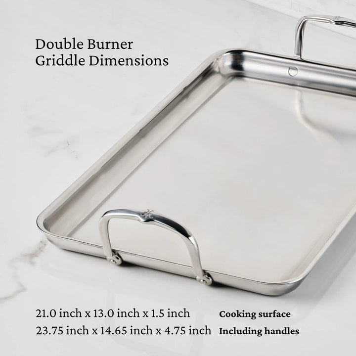 The Hestan Provisions Double Burner Griddle (21 x 13) rests on a white marble surface, highlighting premium stainless steel construction for even heat distribution. Dimensions: 21 x 13 x 1.5 in (cooking), 23.75 x 14.65 x 4.75 in (with handles).