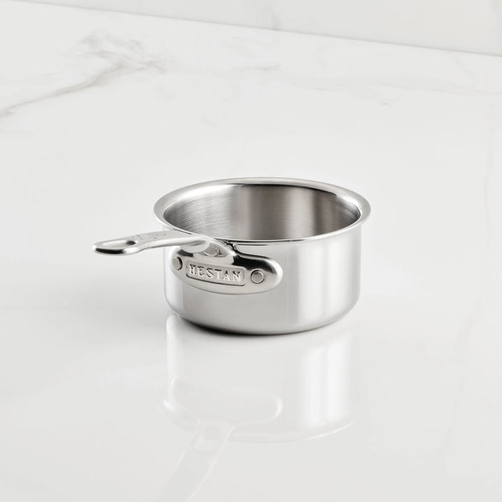 A small stainless steel saucepan from the Hestan Provisions Host and Roast 8-piece set rests empty on a white marble surface, with the brand name engraved on its short handle.