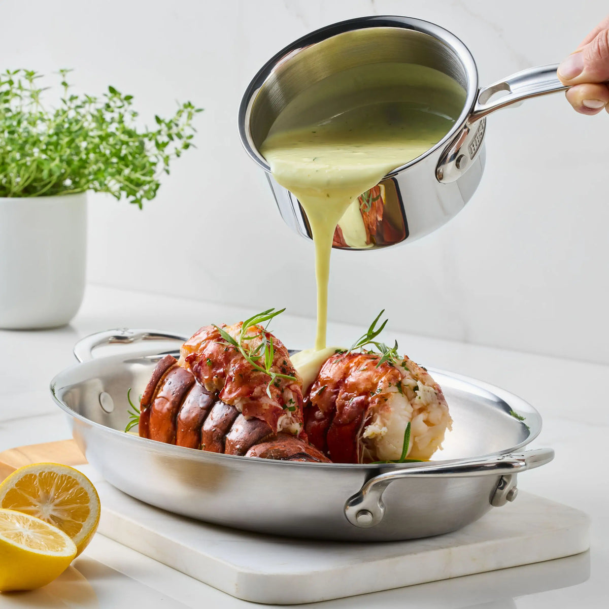 A hand pours creamy sauce from the ProBond® Luxe™ Polished Clad Stainless Steel Butter Warmer, 0.75-Quart, over two lobster tails garnished with fresh herbs; a halved lemon and a potted plant sit nearby on the counter.