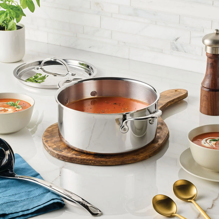 A ProBond® Luxe™ Polished Clad Stainless Steel Soup Pot with Lid, filled with tomato soup, sits on a wooden board beside a bowl of soup and utensils on a white kitchen countertop—highlighting Italian craftsmanship.