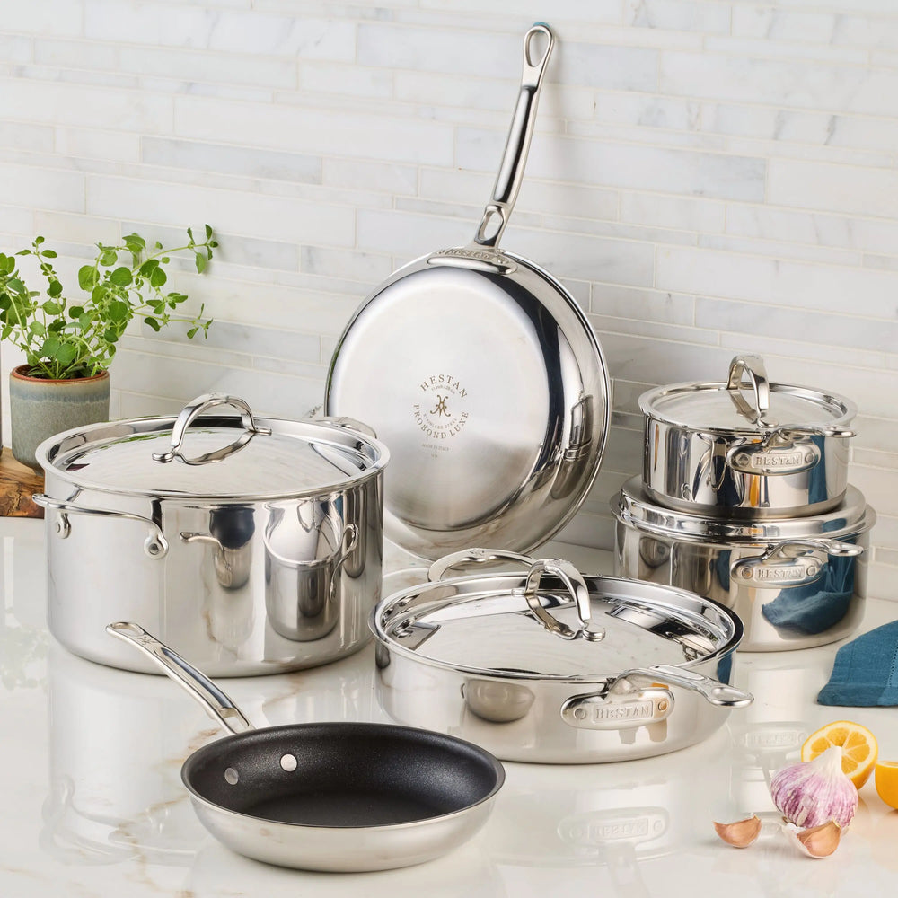 The ProBond® Luxe™ Polished Clad Stainless Steel Premiere Set, 10-Piece, known for Italian craftsmanship, sits on a kitchen counter with a plant, lemon slice, garlic cloves, and blue towel beside the gleaming pots and pans.
