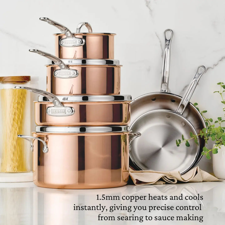 A stunning CopperBond® Aura™ Polished Copper 10-Piece Cookware Set with silver handles is neatly stacked on the countertop, highlighting its blend of style and function. Two frying pans from the same collection hang nearby, enhancing the kitchen’s elegant atmosphere. Next to the gleaming pots and pans are a bundle of dry spaghetti and a potted plant. Text below describes CopperBond® Aura™’s exceptional heat control, making it perfect for precise cooking.