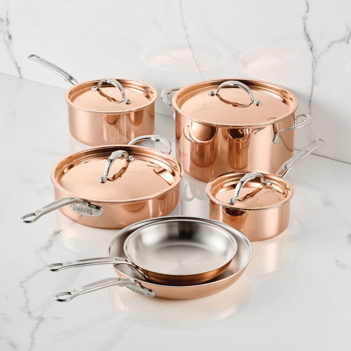 The CopperBond® Aura™ Polished Copper 10-Piece Cookware Set, featuring stainless steel interiors, three lidded pots, and two frying pans, is displayed on a white marble surface with a gray veined background.