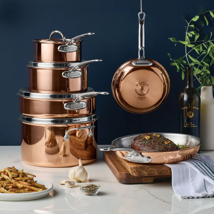 Elevate your kitchen with the elegance of the CopperBond® Aura™ Polished Copper 10-Piece Cookware Set, featuring four nested pots with lids, a frying pan artistically presented on a wooden board with food, and a hanging pan. Complete this professional-grade copper display with accents like a wine bottle, greenery in a vase, garlic bulbs, a spice bowl, and beautifully arranged fries on a pristine white surface.