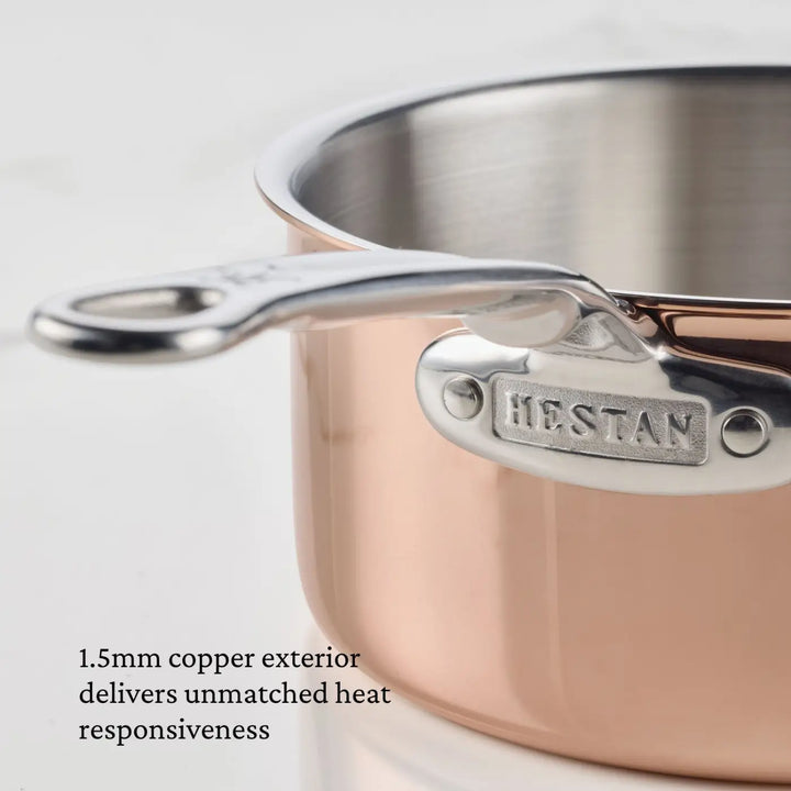 A close-up of a CopperBond® Aura™ Polished Copper 10-Piece Cookware Set saucepan features a gleaming copper exterior and stainless steel handle. Text reads, 1.5mm copper exterior delivers unmatched heat responsiveness.