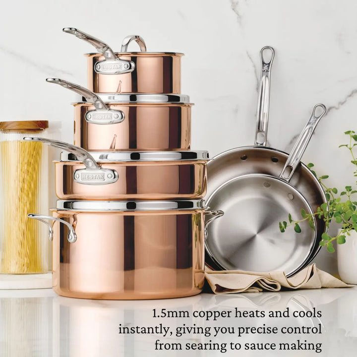 A CopperBond Aura™ Polished Copper 0.75-Quart Butter Warmer sits stacked among a set of professional-grade cookware on a white countertop, with two stainless steel pans propped behind it. Beside the copper cookware, a glass jar filled with pasta and a small plant add kitchen charm. At the bottom, text highlights the superior heating properties of copper.