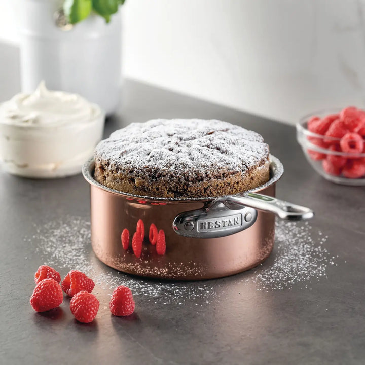 A baked cake dusted with powdered sugar sits elegantly in a CopperBond Aura™ Polished Copper Saucepan with Lid on a gray countertop. Fresh raspberries are scattered nearby, with a bowl of raspberries and a bowl of whipped cream in the background, showcasing the allure of premium copper cookware.