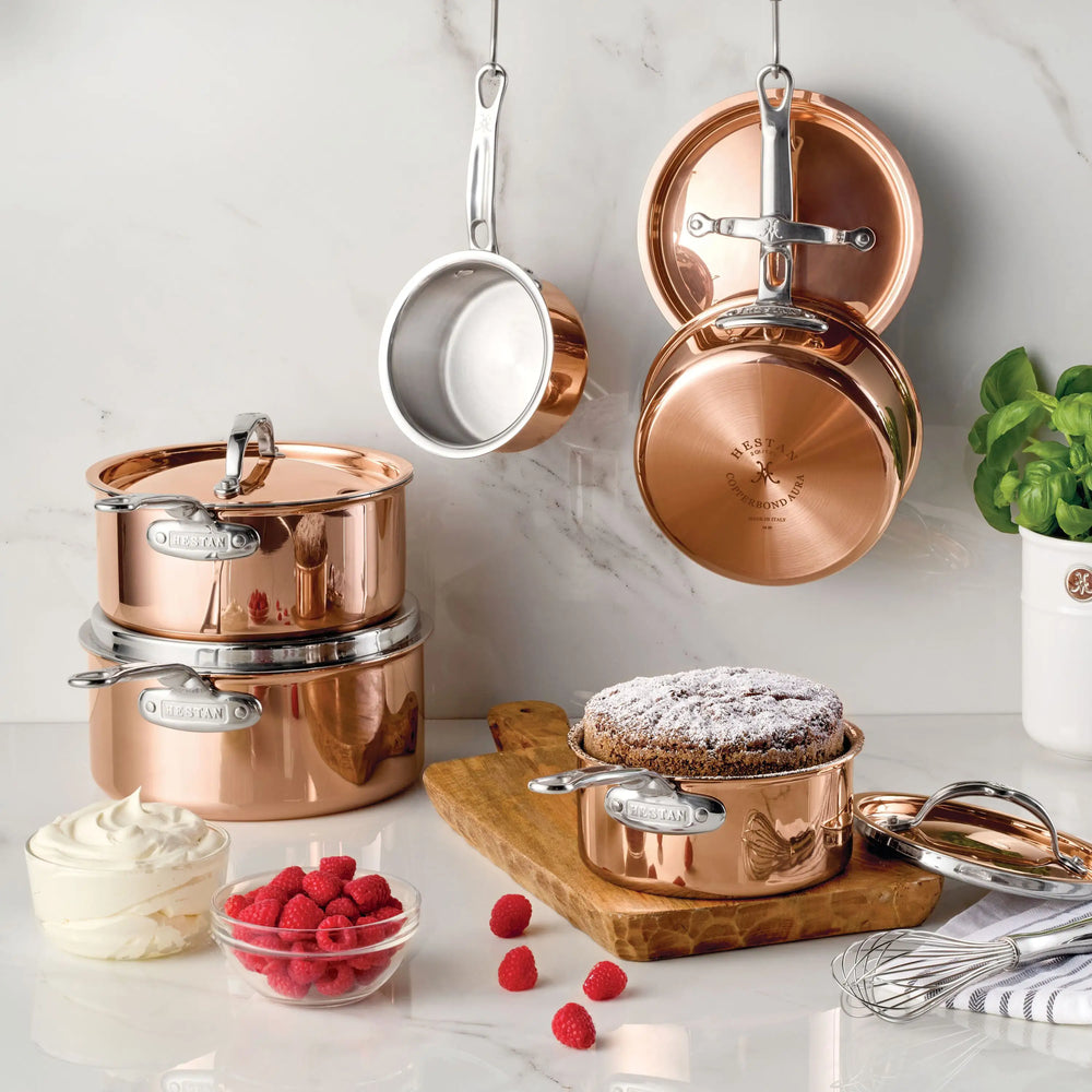 Polished Copper Saucepans with Lid from CopperBond Aura™ abound, stacked and hanging on a marble kitchen wall. A cake in a gleaming saucepan sits on a wooden board near fresh raspberries, whipped cream, a whisk, and a basil plant—showcasing the elegance of CopperBond Aura™ in every detail.