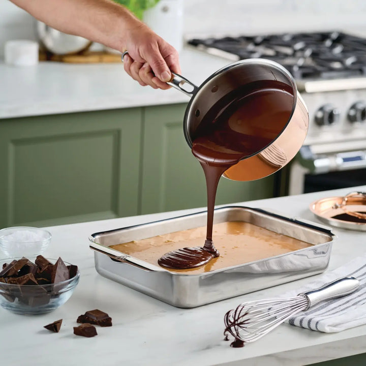 A hand pours melted chocolate from a CopperBond Aura™ Polished Copper Saucepan with Lid onto a layer of light-colored batter in a rectangular metal baking pan. A whisk and chopped chocolate are nearby on the white kitchen counter.