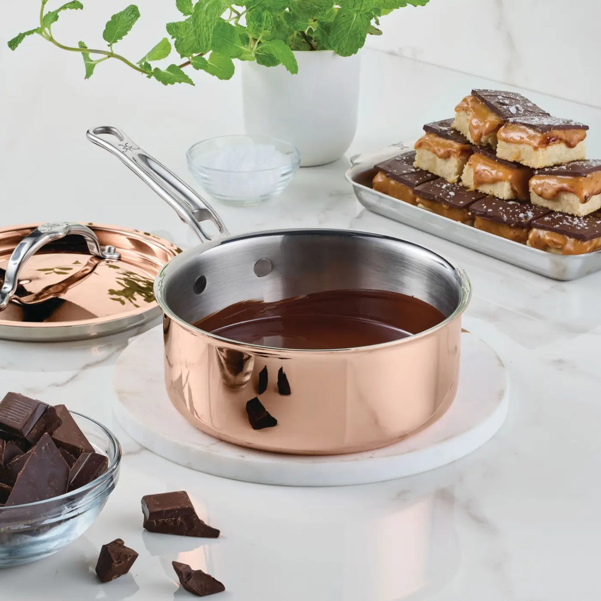 A CopperBond Aura™ Polished Copper Saucepan with Lid filled with melted chocolate sits on a white marble surface. Nearby are a bowl of chocolate pieces, a tray of caramel bars, a pot of mint, a bowl of salt, and the matching gleaming copper lid.