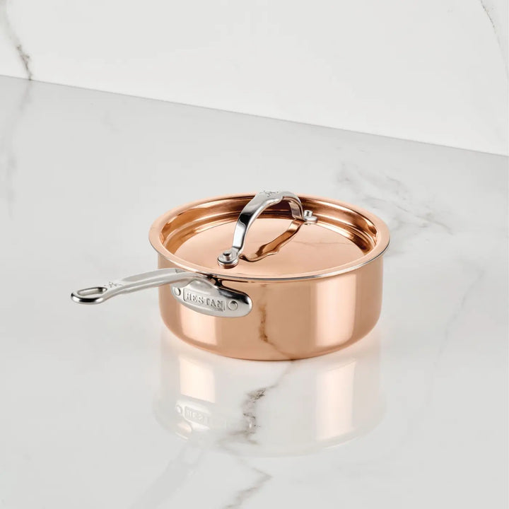 A CopperBond Aura™ Polished Copper Saucepan with Lid, featuring a stainless steel handle and a slightly domed lid with a central handle, sits on a white marble surface with gray veining. A label appears on the side near the handle, highlighting the refined elegance characteristic of premium copper cookware.