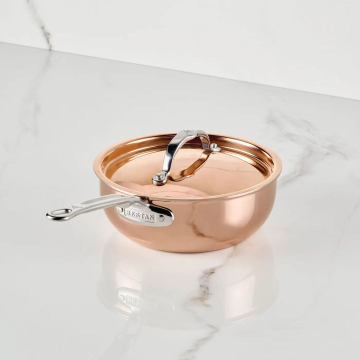 A Polished Copper 2-Quart Saucier with Lid from CopperBond Aura™ rests on a white marble surface. Showcasing the sophistication of professional cookware, both the sleek silver handle and matching lid feature engraved CopperBond Aura™ branding, while the lustrous copper body beautifully reflects ambient light.