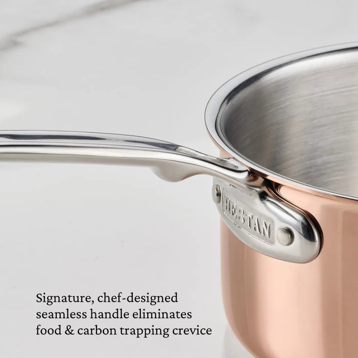 A close-up view of the CopperBond Aura™ Polished Copper 2-Quart Saucier with Lid shows its stainless steel, seamless handle, which features a Hestan label at the base. Text beside the saucier highlights that the seamless design prevents food and carbon from accumulating in crevices, making this piece of copper cookware perfect for both home cooks and those seeking professional-grade performance.