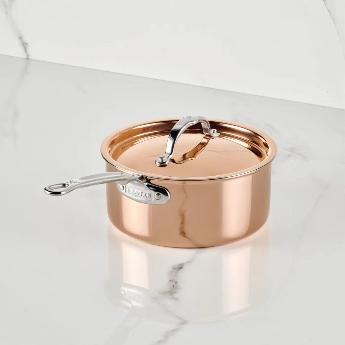 A CopperBond Aura™ Polished Copper Saucepan with Lid, featuring a silver handle, rests on a white marble surface with grey veining and is set against a coordinating marble background. This premium saucepan from the CopperBond Aura™ collection has a shiny, reflective finish and comes complete with a matching handled lid.