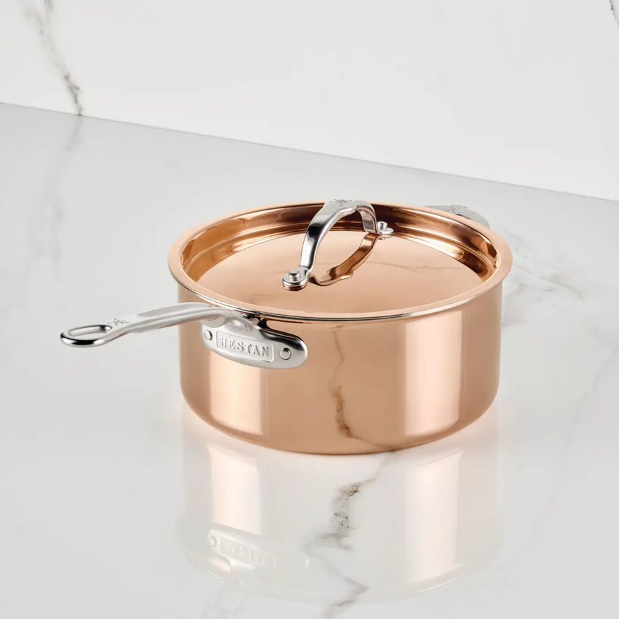 A CopperBond Aura™ Polished Copper Saucepan with Lid sits on a white marble countertop adorned with subtle gray veining. The saucepan, featuring a shiny, reflective surface and an upright lid handle, represents premium copper cookware.