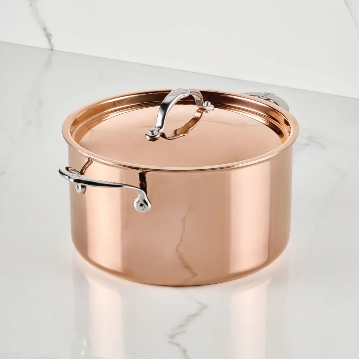 A CopperBond Aura™ Polished Copper Stock Pot with Lid sits on a white marble surface with gray veining. This professional copper cookware piece features a copper-colored body, silver side handles, and a matching lid with a looped handle. Designed for durability and optimal heat retention, it boasts a stainless steel interior.