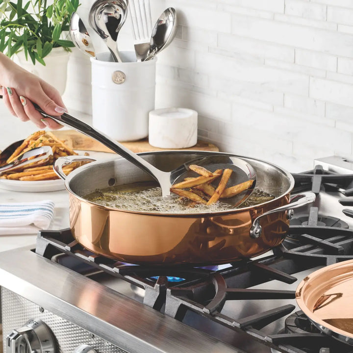 A hand uses a slotted spoon to lift French fries from hot oil in the CopperBond Aura™ Polished Copper Rondeaus with Lid on a gas stove. Cooked fries rest on a plate nearby, while kitchen utensils sit in a container on the counter in the background.