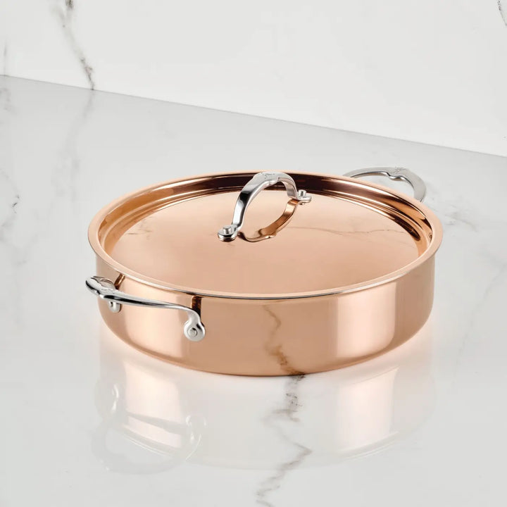 Introducing the CopperBond Aura™ Polished Copper Rondeaus with Lid—a professional-grade, Italian-crafted copper pot featuring a round silhouette and lustrous, reflective finish. Silver handles and an upright lid handle accentuate its elegance as it rests gracefully on a white marble surface, exemplifying premium copper cookware for your kitchen.