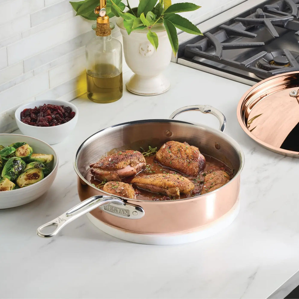 A CopperBond Aura™ Polished Copper Saute Pan with Lid, featuring a stainless steel interior, holds four seasoned pieces of chicken as it rests on a white countertop near a bowl of Brussels sprouts, a bowl of cranberries, olive oil, a potted plant, and a stovetop.