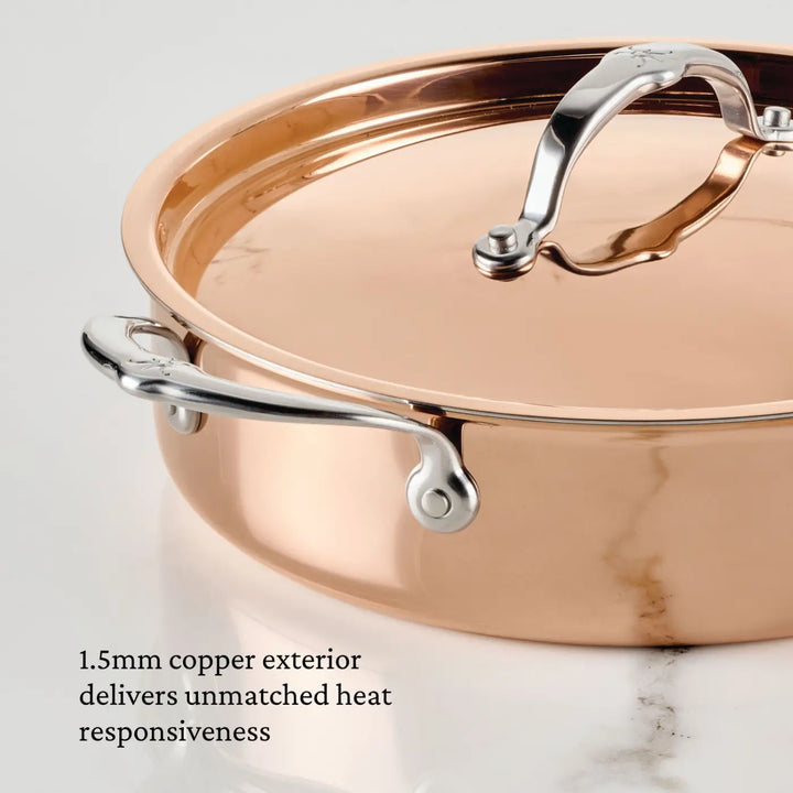 A Polished Copper Saute Pan with Lid from CopperBond Aura™ is displayed on a light marble surface. Featuring a lustrous 1.5mm copper exterior for exceptional heat responsiveness and a robust stainless steel interior, this elegant cookware seamlessly blends high-performance cooking with timeless style.