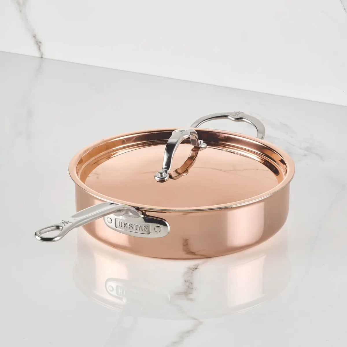 A CopperBond Aura™ Polished Copper Saute Pan with Lid rests on a white marble surface. This premium copper cookware features an arched lid handle and stainless steel handles, with the brand name elegantly marked on the handle. The gleaming stainless steel interior reflects the light, set against a backdrop of a white marble wall.