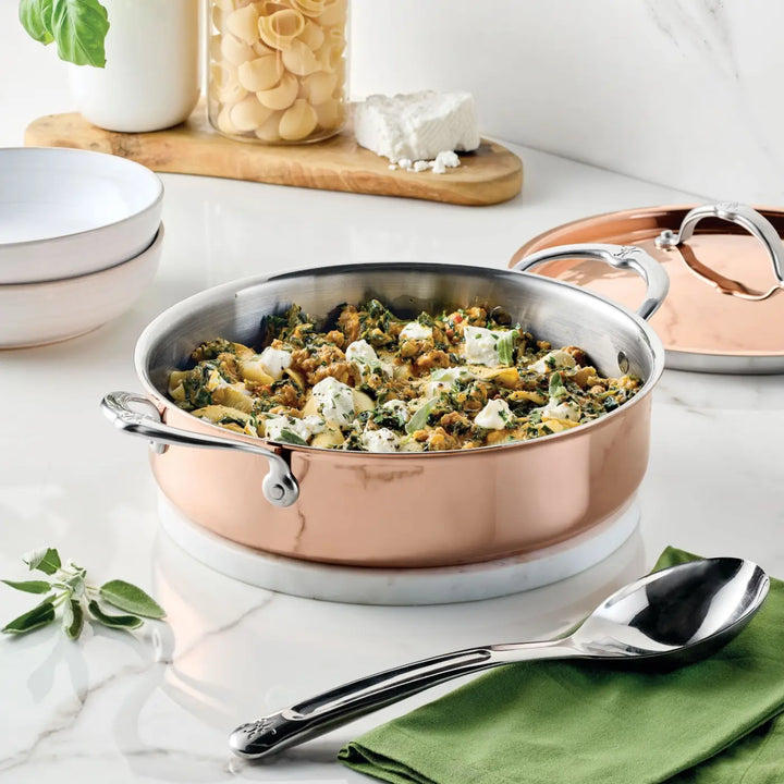 A CopperBond Aura™ Polished Copper Rondeaus with Lid, crafted in the style of professional-grade cookware and featuring sturdy handles, is filled with a vegetable and cheese dish. Nearby are a serving spoon, a green napkin, and two white bowls. Behind the rondeau sit a glass jar of pasta, a block of cheese, and fresh herbs—perfectly evoking an Italian-crafted kitchen scene.