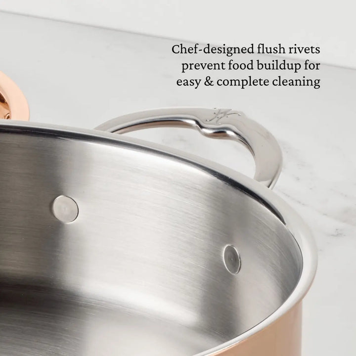 Close-up of a CopperBond Aura™ Polished Copper Rondeaus with Lid, featuring professional-grade stainless steel construction and chef-designed flush rivets on the inside wall. Part of the handle and a marble countertop are also visible. Text highlights how the Italian-crafted cookware’s innovative flush rivets prevent food buildup for easier cleaning, offering both elegance and practicality.