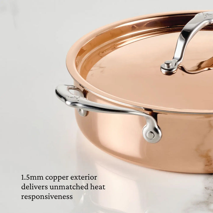 A close-up of the CopperBond Aura™ Polished Copper Rondeaus with Lid, Italian crafted and featuring stainless steel handles, sits on a white marble surface. Text reads: 1.5mm copper exterior delivers unmatched heat responsiveness.