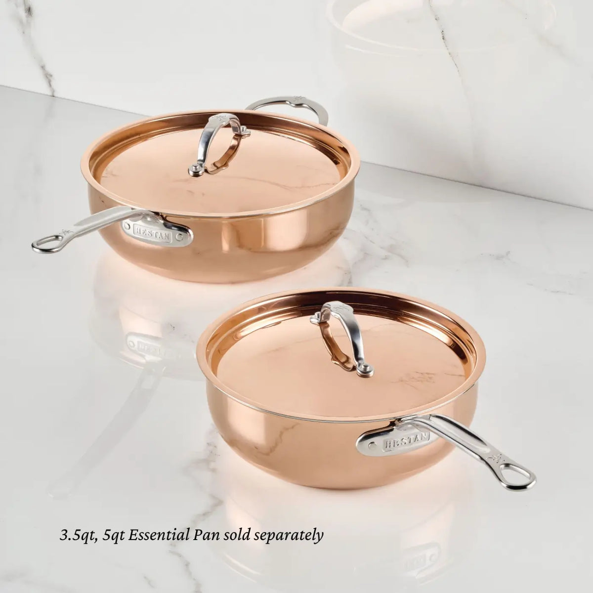 Two Italian-crafted Polished Copper Essential Pans with Lids from CopperBond Aura™ are displayed on a white marble surface. Each copper-colored, professional-grade pan features a metal label and elegant silver handles, while the lids are topped with curved handles. A note below indicates: 3.5qt and 5qt sizes sold separately.