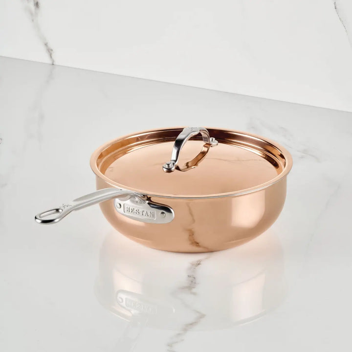 A CopperBond Aura™ Polished Copper Essential Pan with Lid rests on a white marble surface streaked with gray veins. Expertly Italian-crafted and professional-grade, this striking copper cookware showcases the brand name engraved along the side of its silver handle.