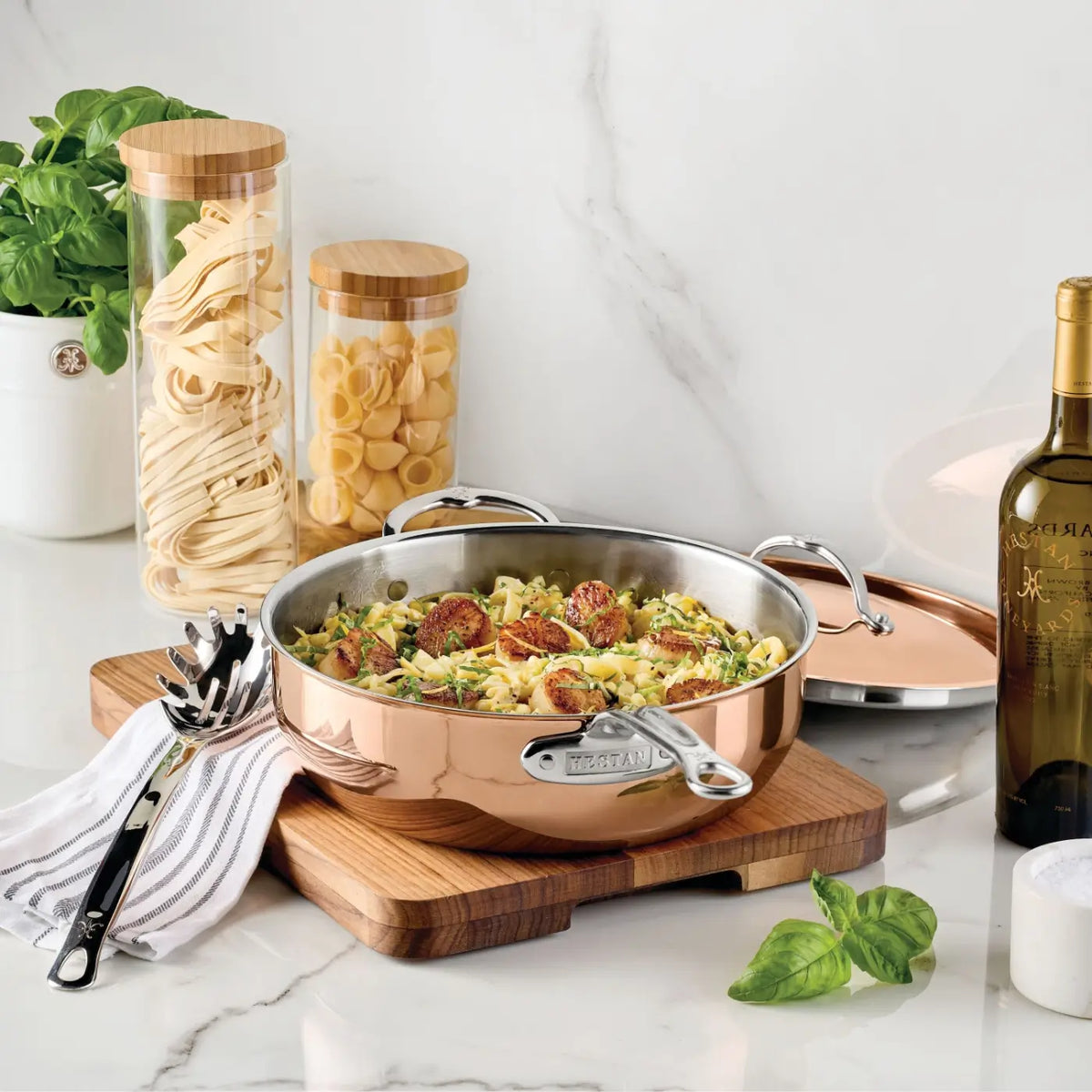 A Polished Copper Essential Pan with Lid from the CopperBond Aura™ collection—an Italian-crafted, professional-grade pan—holds pasta and vegetables as it rests on a wooden board. Surrounding it are a wine bottle, jars of pasta, a utensil atop a striped towel, a sprig of basil, and a white container, all elegantly arranged against a white marble background.