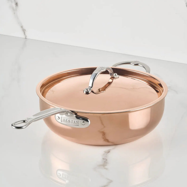 A polished copper saucepan with a lid and two metal handles from CopperBond Aura™ is placed on a white marble countertop. This expertly crafted pan gleams with a reflective finish, featuring the CopperBond Aura™ brand on the side handle—an exquisite example of professional-grade Polished Copper Essential Pans with Lid.
