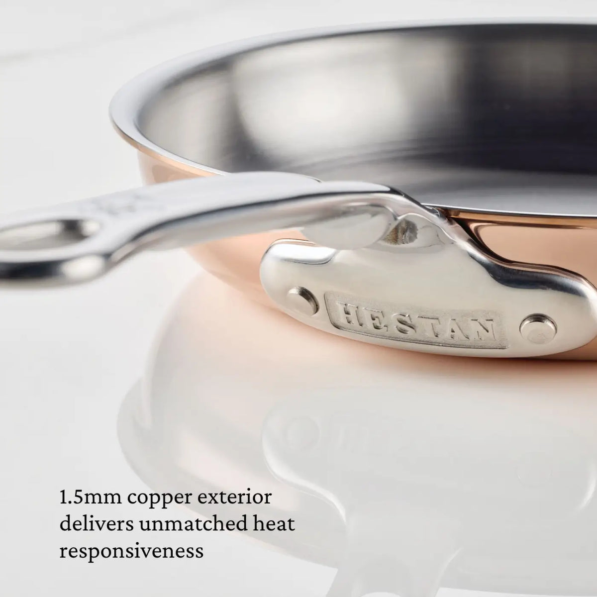 Close-up of the CopperBond Aura™ Polished Copper Skillets’ stainless steel handle and copper rim, placed on a white surface. This premium piece features text that reads: 1.5mm copper exterior delivers unmatched heat responsiveness.