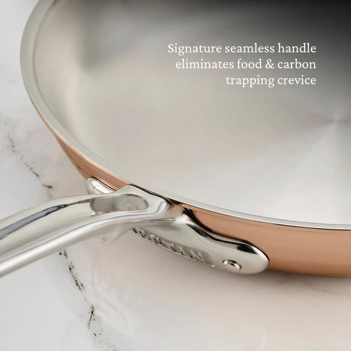 Close-up of a Polished Copper Skillet from the CopperBond Aura™ collection, featuring a seamless metal handle. Text on the skillet reads: Signature seamless handle eliminates food & carbon trapping crevice. The copper skillet rests elegantly on a marble surface.