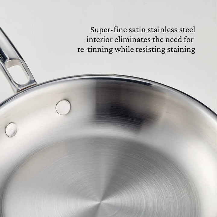Close-up of a polished copper skillet with a curved handle, part of the CopperBond Aura™ collection. Text beside the pan reads: Super-fine satin stainless steel interior eliminates the need for re-tinning while resisting staining, showcasing the exceptional quality found in premium copper cookware.