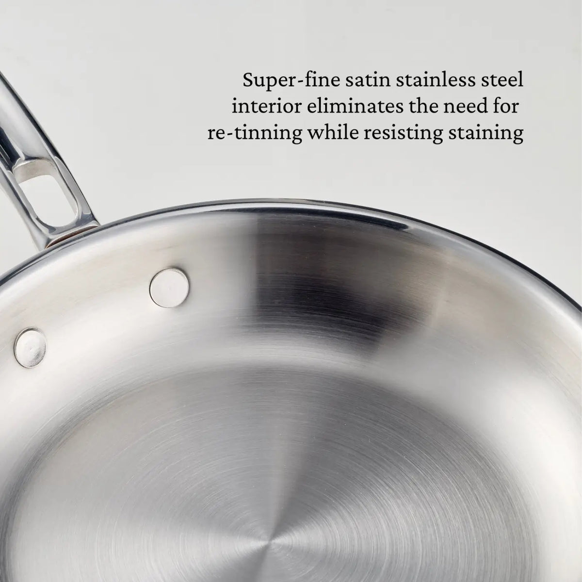 Close-up of a polished copper skillet with a curved handle, part of the CopperBond Aura™ collection. Text beside the pan reads: Super-fine satin stainless steel interior eliminates the need for re-tinning while resisting staining, showcasing the exceptional quality found in premium copper cookware.