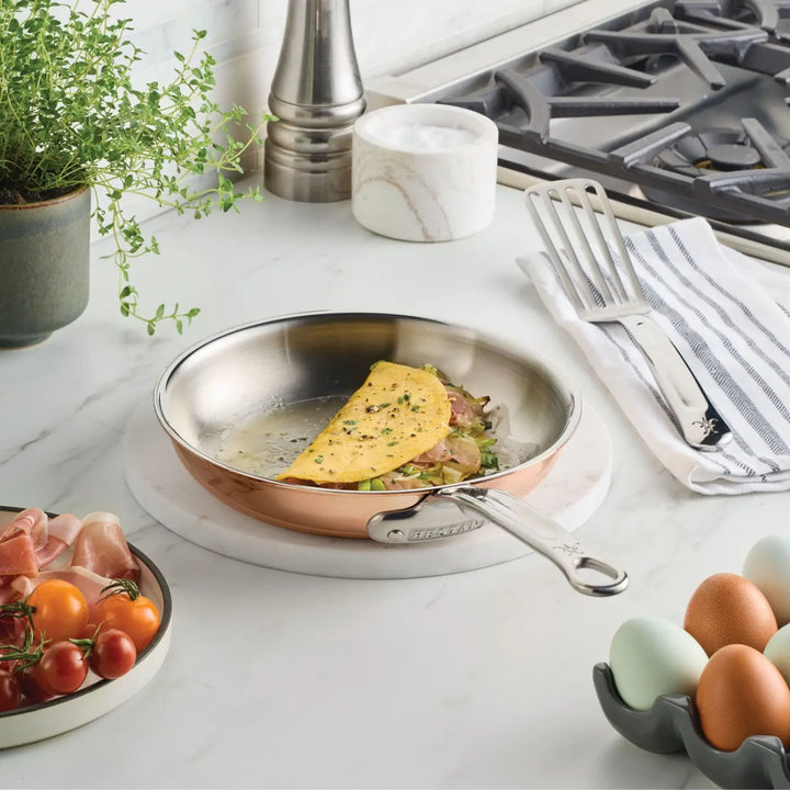 A half-folded omelet with herbs sits in a gleaming CopperBond Aura™ Polished Copper Skillet on a marble countertop. Nearby are cherry tomatoes, prosciutto, eggs, a striped towel with a spatula, a potted plant, and a stove.