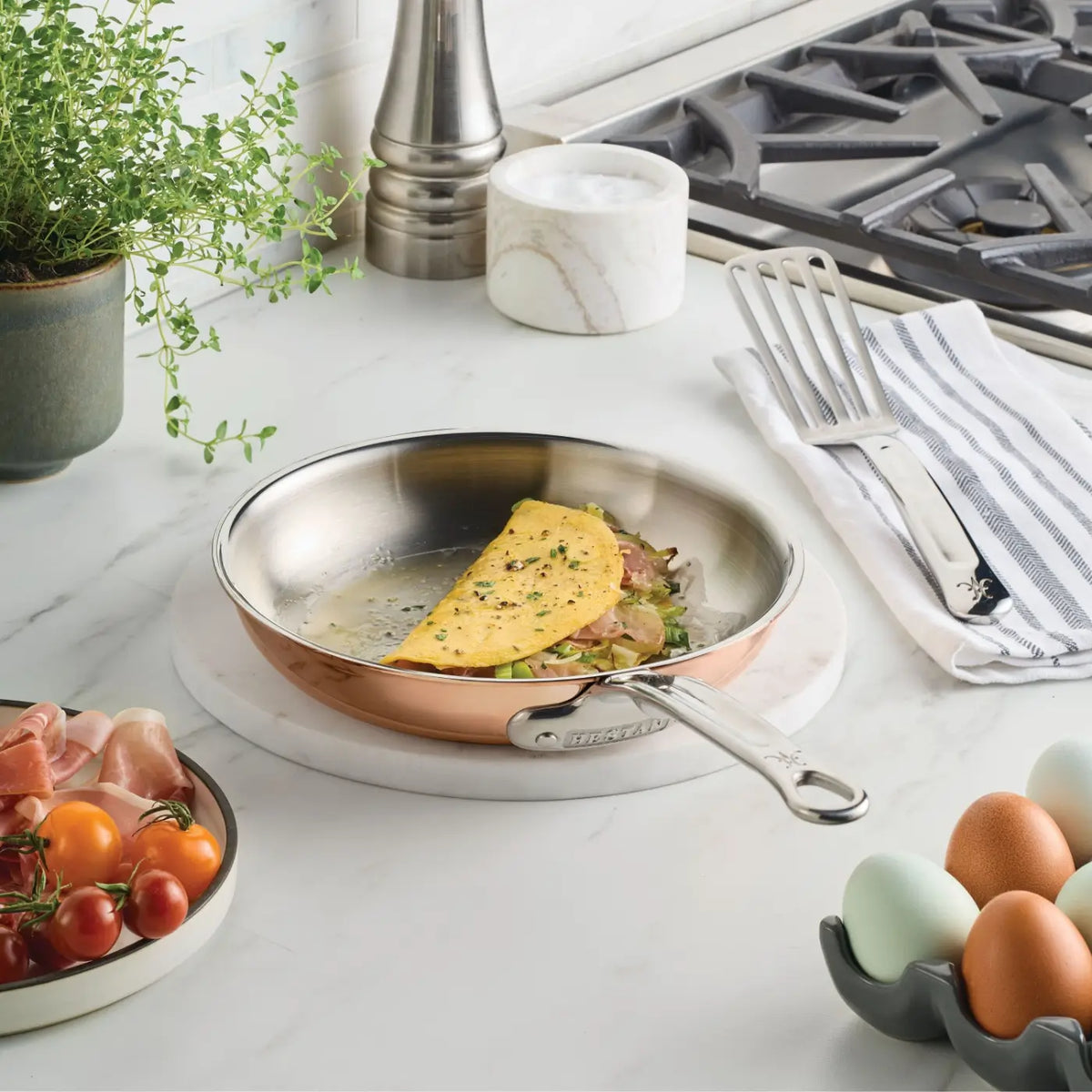 A half-folded omelet with herbs sits in a gleaming CopperBond Aura™ Polished Copper Skillet on a marble countertop. Nearby are cherry tomatoes, prosciutto, eggs, a striped towel with a spatula, a potted plant, and a stove.