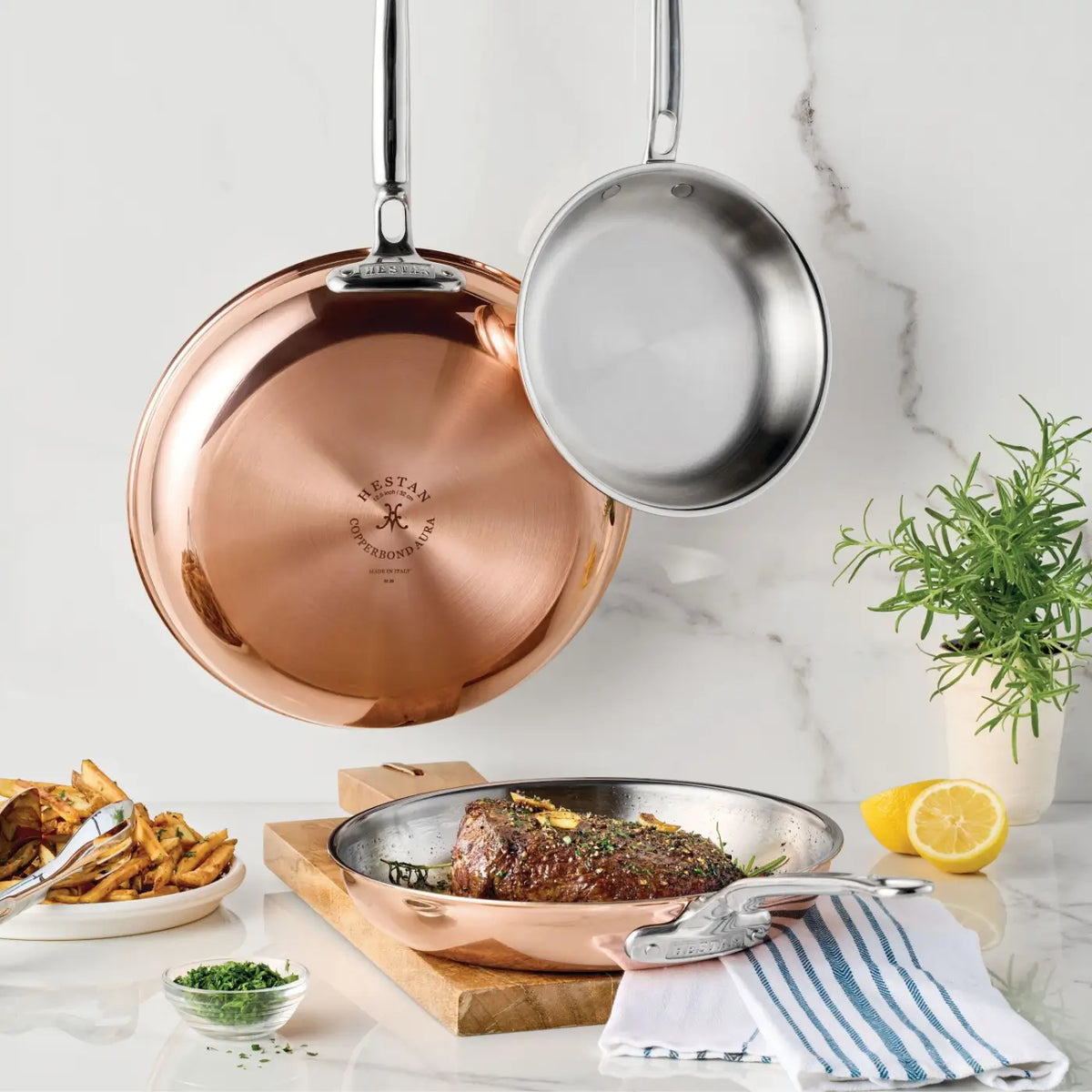 Two CopperBond Aura™ Polished Copper Skillets and a stainless steel pan are hanging on a white wall. A third skillet on the counter holds a cooked steak with herbs. French fries, a fork, a lemon half, and a potted herb sit nearby. A striped towel lies at the bottom right.