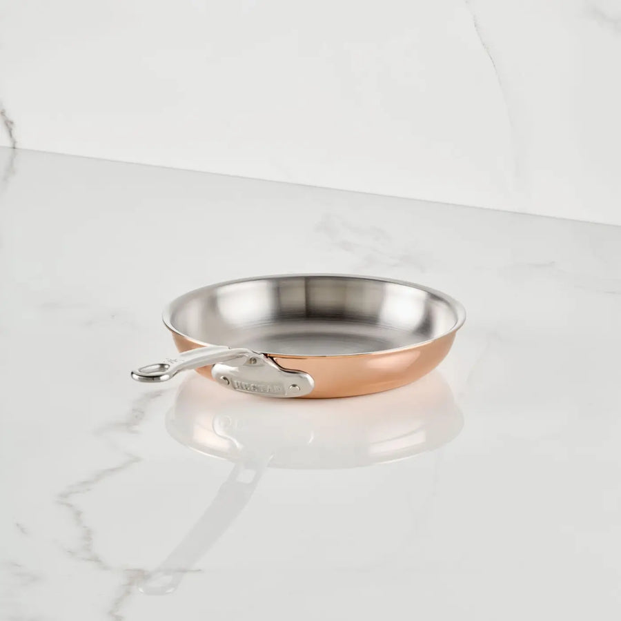 A CopperBond Aura™ Polished Copper Skillet with a stainless steel interior and metal handle sits elegantly on a white marble surface against a matching white marble background.