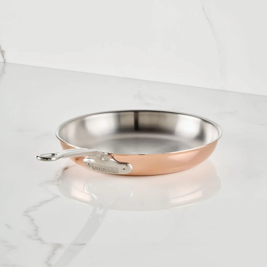 A CopperBond Aura™ Polished Copper Skillet, showcasing a stainless steel interior, gleaming copper exterior, and a polished metal handle, rests on a white marble surface in front of an elegant white marble wall.